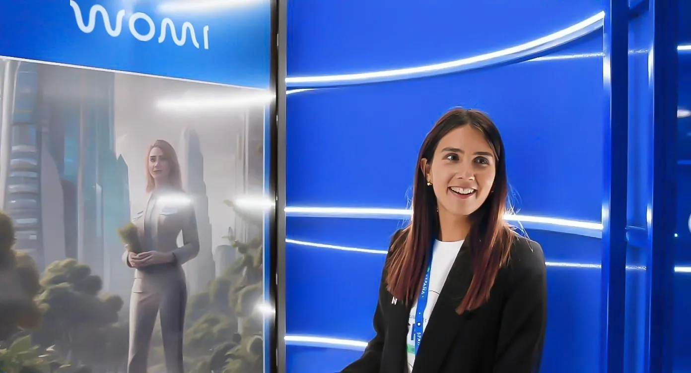 Smiling woman beside the WOMI booth, whose screen shows an AI-transformed photo in a futuristic cityscape